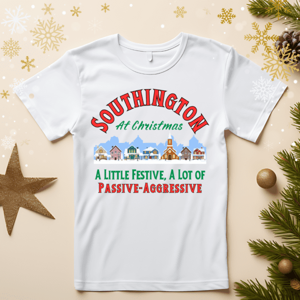 Festive T-shirt mockup featuring a holiday design with the text 'Southington At Christmas' above an illustration of a charming winter village. The tagline reads, 'A Little Festive, A Lot of Passive-Aggressive,' in bold red and green typography. The T-shirt is displayed with holiday-themed decor, including snowflakes, pine branches, and golden stars, evoking a cheerful Christmas atmosphere.